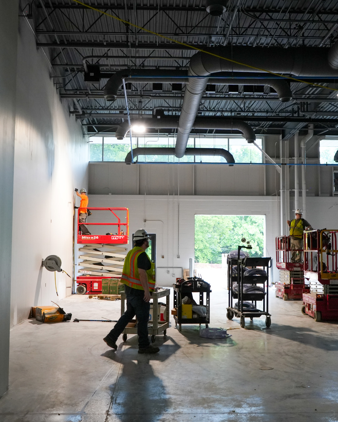 Construction crews work from scissor lifts inside a TCAT Jacksboro high-bay classroom as wall finishes and overhead mechanical systems are completed.